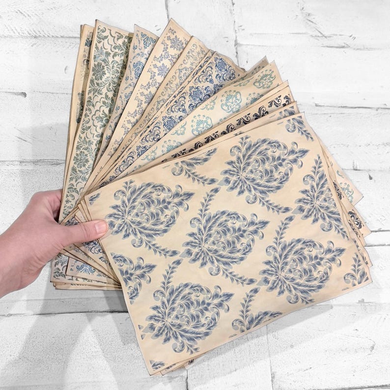May include: A collection of vintage-style paper sheets with various blue floral and ornamental patterns. The papers are held in a fan shape, showcasing the different designs on a cream-colored background. The papers have a distressed, aged appearance.