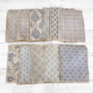 May include: A collection of rectangular paper sheets with various blue and off-white floral and damask patterns. The sheets are arranged on a white wooden surface, showcasing a variety of vintage-inspired designs. The patterns include stylized flowers and geometric shapes.