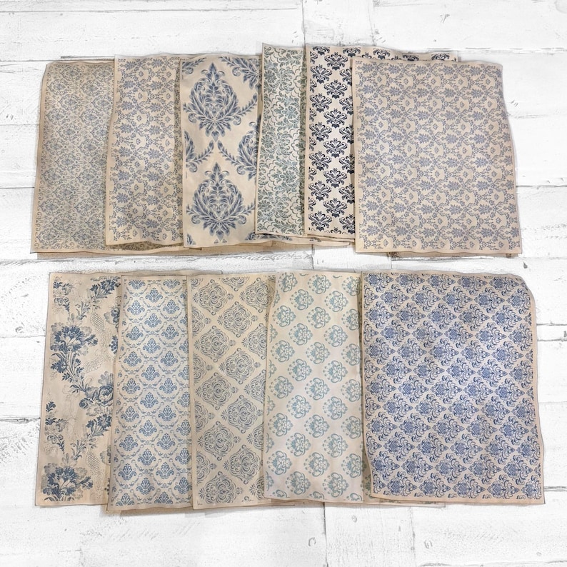 May include: A collection of rectangular paper sheets with various blue and off-white floral and damask patterns. The sheets are arranged on a white wooden surface, showcasing a variety of vintage-inspired designs. The patterns include stylized flowers and geometric shapes.
