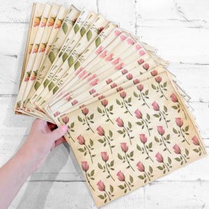 May include: A collection of vintage-style paper sheets with floral designs. The sheets feature pink roses and green leaves on a cream-coloured background. Some sheets have a repeating rose pattern, while others have a variety of floral and leaf patterns.