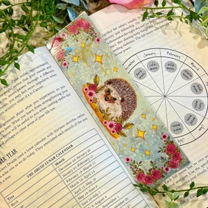 May include: A bookmark featuring a watercolor illustration of a hedgehog with pink flowers and stars on a blue background. The bookmark also includes a circular chart with the names of the months of the year and the phases of the moon.