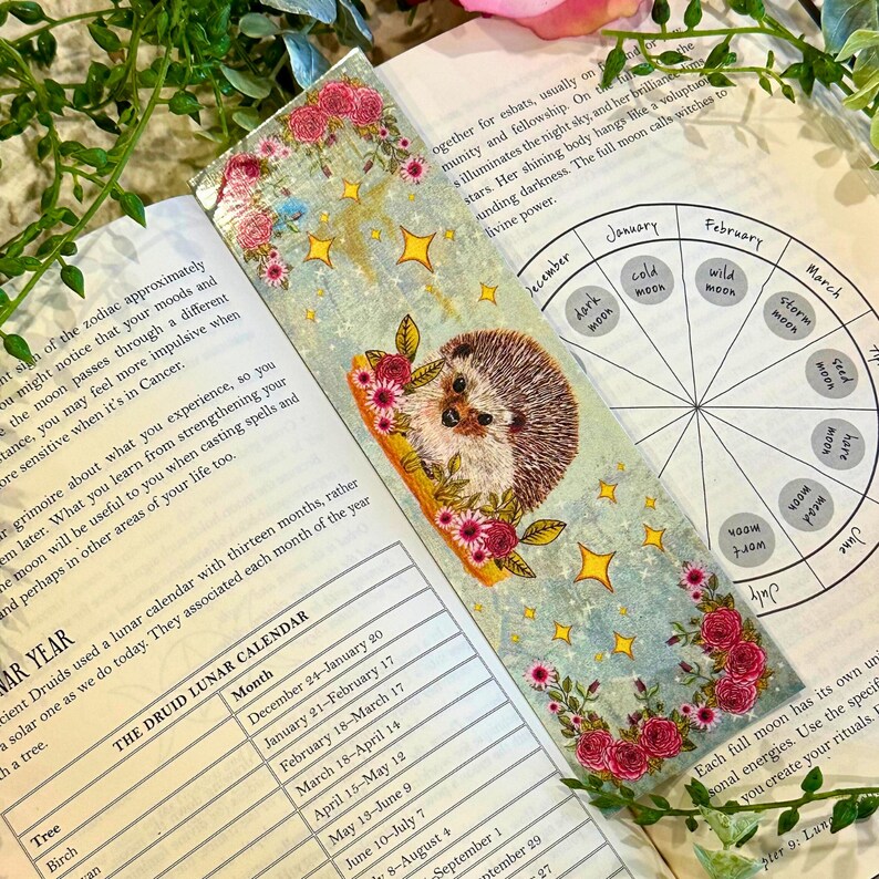 May include: A bookmark featuring a watercolor illustration of a hedgehog with pink flowers and stars on a blue background. The bookmark also includes a circular chart with the names of the months of the year and the phases of the moon.