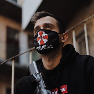 Op de afbeelding: Zwart gezichtsmasker met het Umbrella Corporation-logo, een rood-wit embleem op een zwarte achtergrond. De persoon draagt een zwarte hoodie met een rood-wit logo en houdt een zilveren geweer vast.