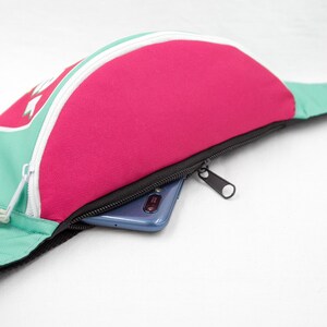 May include: A fanny pack in a vibrant color block design. The bag features a pink center panel, teal side panels, and a white zipper. A black strap and zipper pull complete the design. A smartphone is visible inside the zippered pocket.