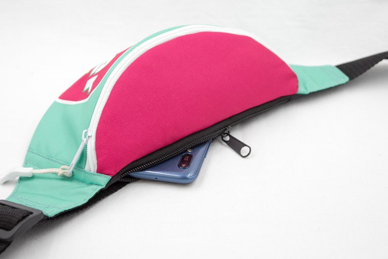 May include: A fanny pack in a vibrant color block design. The bag features a pink center panel, teal side panels, and a white zipper. A black strap and zipper pull complete the design. A smartphone is visible inside the zippered pocket.