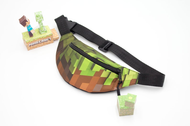 May include: A green and brown Minecraft-themed fanny pack with a black adjustable strap and a zipper closure. The fanny pack has a pixelated design.