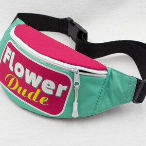 May include: A teal fanny pack with a pink top section and a white zipper. The pack features the words "Flower Dude" in white and yellow lettering on a pink and white label. The strap is black.