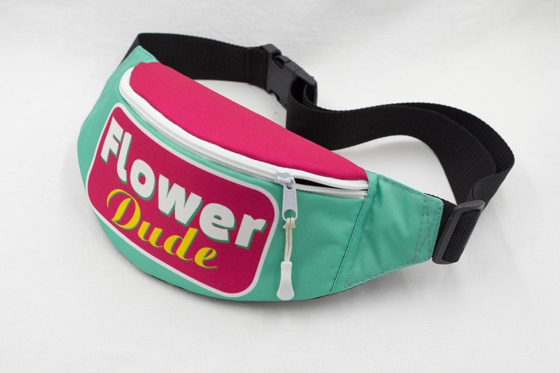 May include: A teal fanny pack with a pink top section and a white zipper. The pack features the words "Flower Dude" in white and yellow lettering on a pink and white label. The strap is black.