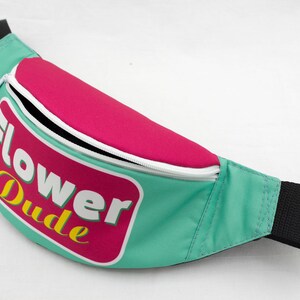 May include: A teal fanny pack with a pink top section and a white zipper. The pack features the text "Flower Dude" in a pink and white rectangular design with yellow accents. The strap is black.