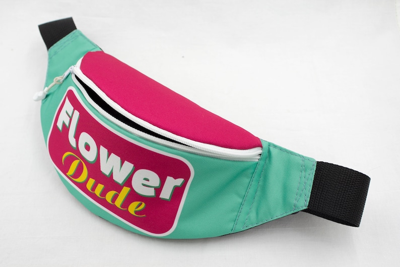 May include: A teal fanny pack with a pink top section and a white zipper. The pack features the text "Flower Dude" in a pink and white rectangular design with yellow accents. The strap is black.