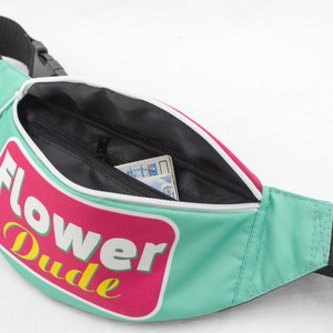May include: A teal fanny pack with a black strap and zipper. The pack features a pink and white graphic with the words "Flower Dude" in yellow and white. The interior is black, and a card is visible inside.