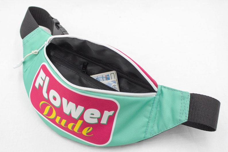 May include: A teal fanny pack with a black strap and zipper. The pack features a pink and white graphic with the words "Flower Dude" in yellow and white. The interior is black, and a card is visible inside.