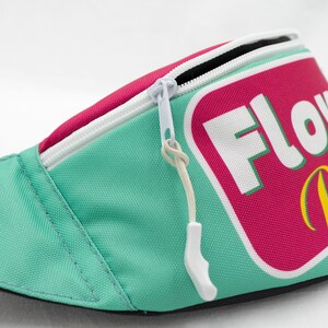 May include: A teal fanny pack with a white zipper and a pink and white rectangular patch that reads "FLOW Dude" in white and yellow lettering. The fanny pack has a white zipper pull.