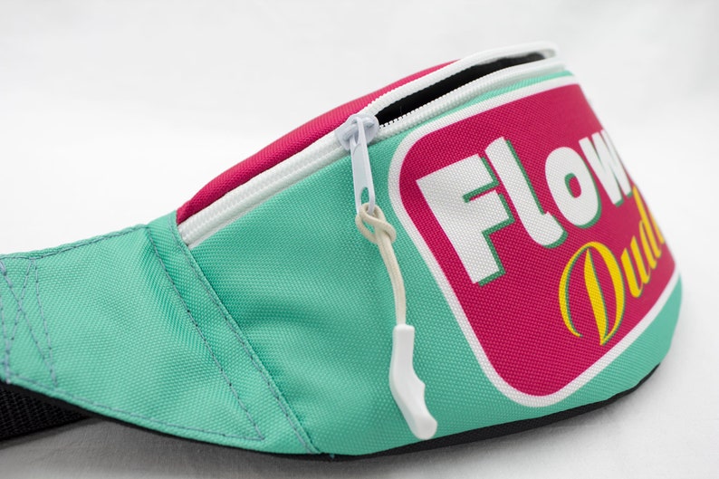 May include: A teal fanny pack with a white zipper and a pink and white rectangular patch that reads "FLOW Dude" in white and yellow lettering. The fanny pack has a white zipper pull.