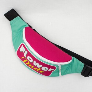May include: A teal, pink, and black fanny pack with the words "Flower Dude" in a retro font. The fanny pack has a white zipper and a black adjustable strap. The bag is designed to be worn around the waist.