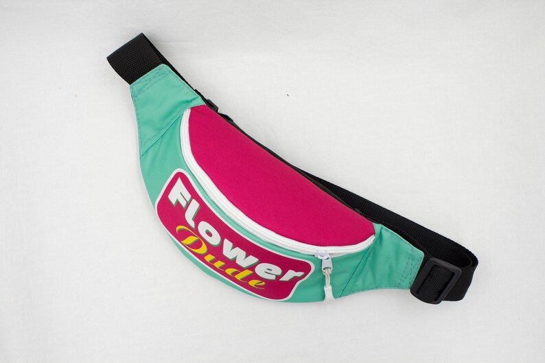 May include: A teal, pink, and black fanny pack with the words "Flower Dude" in a retro font. The fanny pack has a white zipper and a black adjustable strap. The bag is designed to be worn around the waist.