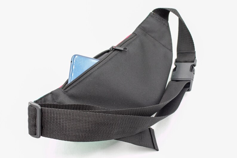 May include: Black fanny pack with a zippered pocket, a black adjustable strap, and a black plastic buckle. A blue smartphone is partially visible inside the pocket. The pack has a colorful accent.