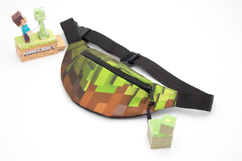 May include: A green and brown fanny pack with a zipper closure and a black adjustable strap. The fanny pack has a Minecraft pixelated design.