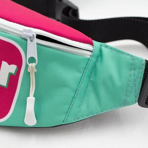 May include: A close-up of a fanny pack with a teal body, a pink top, and a white zipper. The fanny pack has a black adjustable strap and a buckle. The text "Flower Ride" is visible on the front.