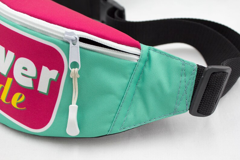 May include: A close-up of a fanny pack with a teal body, a pink top, and a white zipper. The fanny pack has a black adjustable strap and a buckle. The text "Flower Ride" is visible on the front.