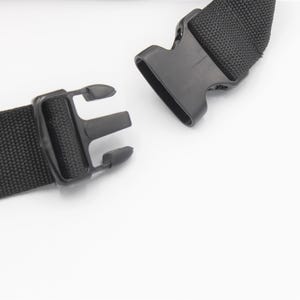 May include: Close-up of a black nylon strap with a plastic buckle. The strap is woven and has a textured appearance. The buckle is open, showing the two parts that connect. The background is white.