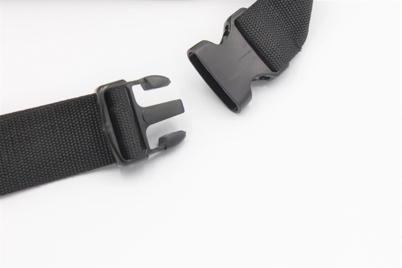 May include: Close-up of a black nylon strap with a plastic buckle. The strap is woven and has a textured appearance. The buckle is open, showing the two parts that connect. The background is white.