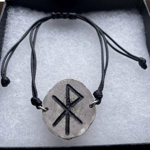 May include: A gray stone pendant with a black rune symbol, strung on a black adjustable cord bracelet. The pendant is oval-shaped and has a textured surface. The bracelet is presented in a black gift box.