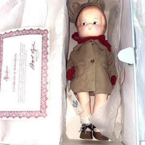 May include: A collectible doll in a box with a certificate of authenticity. The doll has blonde hair, blue eyes, and wears a tan coat, matching hat, red scarf, red mittens, white socks, and brown shoes. The certificate text reads Effanbee Certificate of Authenticity.