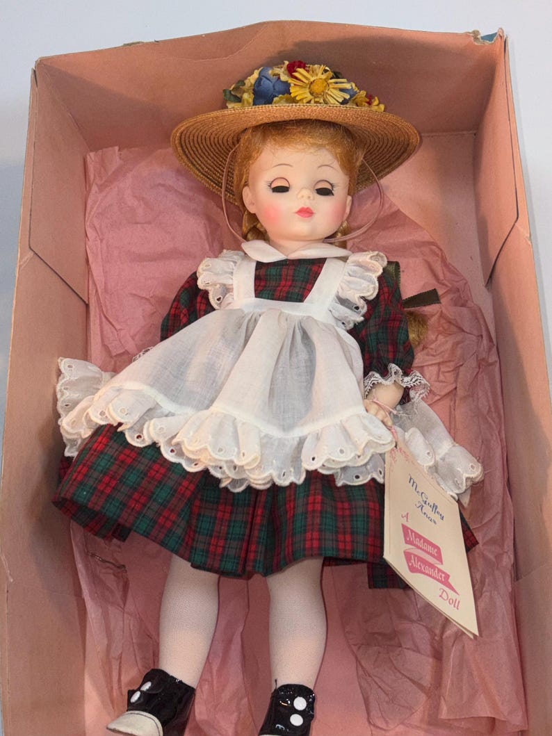 May include: A red and green plaid dress with a white apron and lace trim. The doll is wearing a straw hat with flowers and black shoes with white polka dots. The doll is holding a tag that reads 'McCalls Ann A Madame Alexander Doll.'