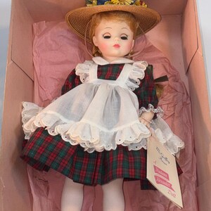 May include: A vintage doll, "M. Guffey Anne," by Madame Alexander, is displayed in its original box. The doll has red hair, a straw hat with floral accents, and wears a plaid dress with a white apron. The doll is wearing black and white shoes.