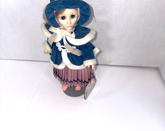 May include: A vintage doll wearing a blue velvet hat and coat with white trim. The doll has a pink pleated skirt and is standing on a gray base. A tag is attached to the base.
