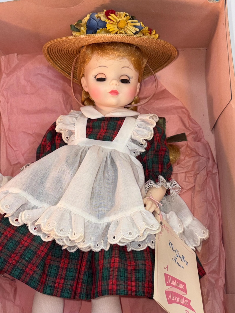 May include: A vintage doll with red hair, wearing a plaid dress with a white apron and a straw hat adorned with flowers. The doll is in a pink box, with a tag that reads "McGuffey Ana" and "Madame Alexander".