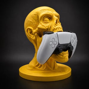 Könnte beinhalten: Ein gelber, schädelförmiger Ständer mit strukturierter, anatomischer Oberfläche und weit geöffnetem Mund, der einen weißen und schwarzen kabellosen Videospiel-Controller hält. Die Basis ist kreisförmig und flach und stützt die Skulptur.