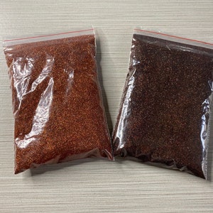 May include: Two clear plastic bags filled with reddish-brown and dark brown granules. The bags are sealed with a red zipper closure. The granules appear to be a fine, granular substance, possibly a spice or a food product.