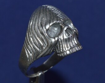 Può includere: Un anello in argento a forma di teschio con un design dettagliato. Il teschio ha una superficie testurizzata e una mascella prominente.