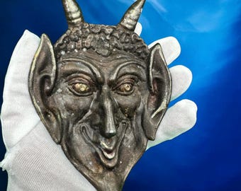 May include: A silver-colored metal mask of a devil with horns, a wide smile, and large ears. The mask is held in a white gloved hand.