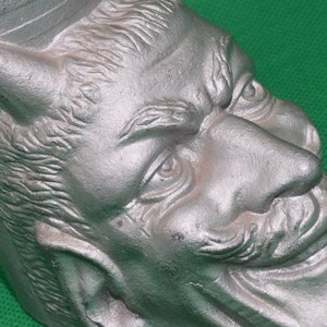 Puede incluir: Una escultura de metal de color plateado de la cara de un diablo con un solo cuerno, el ceño fruncido y una sonrisa traviesa.