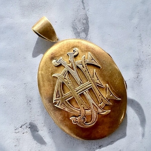 May include: A gold oval locket with a monogram engraved on the front. The locket has a loop at the top for attaching a chain.