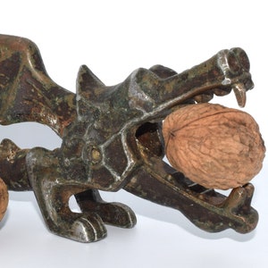 Puede incluir: Un cascanueces de metal vintage en forma de dragón con una nuez en su boca. El dragón está detallado con escamas y tiene un ala en un lado. El cascanueces está pintado en tonos marrones y verdes.