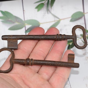May include: Two antique keys with ornate metal handles and rusty brown finishes. The keys are held in a hand against a white wooden background.