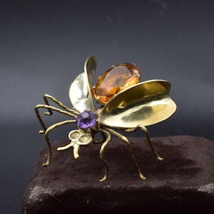 May include: A gold-toned insect brooch with a large amber-colored gemstone on its back and a smaller purple gemstone on its head. The brooch has delicate wire legs and stylized wings.