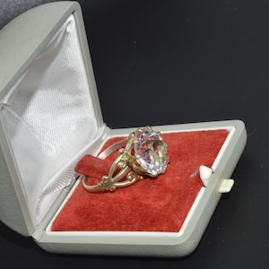 May include: A silver ring with a large clear gemstone set in a decorative filigree design. The ring is displayed in a grey velvet ring box with a red velvet interior.
