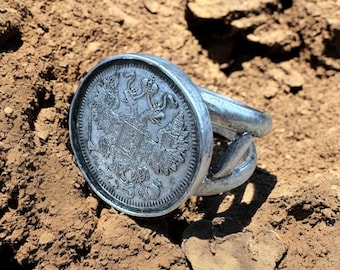 May include: A silver ring with a large coin-shaped face featuring a detailed design of a coat of arms with two eagles and a shield. The ring is lying on a brown dirt surface.