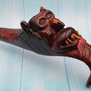 May include: A wooden walking stick carved in the shape of a grotesque face with a large, open mouth and sharp teeth. The stick is painted in shades of brown and red.