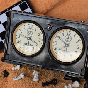 May include: A black chess clock with two round analog faces, each with white hands and black numbers. The clock is sitting on a brown surface with chess pieces scattered around it. A black and white chessboard is visible in the background.