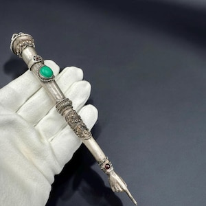 Może przedstawiać: Srebrne pióro z zielonym kamieniem szlachetnym i końcówką w kształcie dłoni. Pióro jest ozdobione misternymi wzorami kwiatowymi i ma zielony kamień szlachetny osadzony w środku.