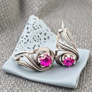 May include: A silver ring and matching earrings set. The jewelry features a bright pink gemstone set in an ornate, swirling design. The ring and earrings are displayed on a light blue surface against a textured background.