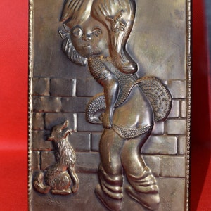 May include: A brass wall plaque with a raised relief design of a girl and a dog. The girl is wearing a dress and has long hair. The dog is sitting next to her. The plaque has a textured background and a decorative border.