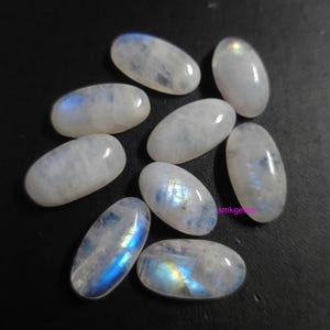Peut inclure: Plusieurs cabochons ovales en pierre de lune. Les pierres sont principalement blanches avec des reflets bleus irisés et arc-en-ciel. Les pierres précieuses polies sont présentées sur un fond sombre, mettant en valeur leur beauté naturelle.