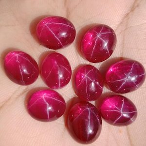 May include: A collection of nine oval-shaped, deep red star rubies. Each gem has a prominent six-ray star pattern, with white lines radiating from the center. The rubies are smooth and polished, reflecting light and set against a warm, skin-toned background.